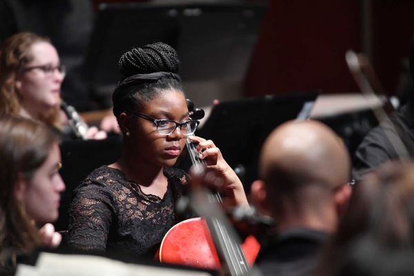 A young woman wearing glasses plays the cello in an orchestra