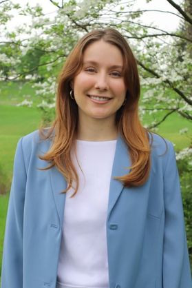 Headshot of WVU Fulbright Scholar Lily Wright. She is standing outside with grass behind her as well as a tree full of white blooms. She is wearing a light blue blazer over a white shirt. She has long, auburn colored hair. 