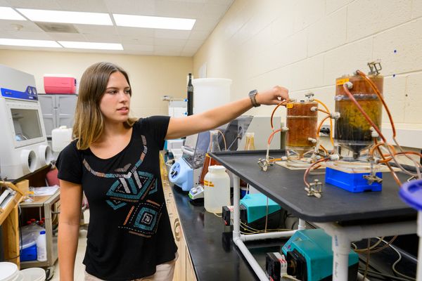 Carley Shingleton, a graduate research assistant at the Statler College is shown here in a lab pointing to two containers of contaminated water. She has shoulder length hair and is wearing a black, short-sleeve top. 