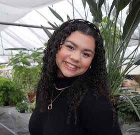 Headshot of WVU student Brogan Dozier. She is pictured in a greenhouse with plants on a table behind her. She has long, curly black hair and is wearing a black turtleneck with a long necklace. 