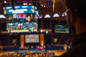 A wide shot is shown of the WVU Coliseum on Commencement Day. In the foreground, a person takes a photo on a phone.