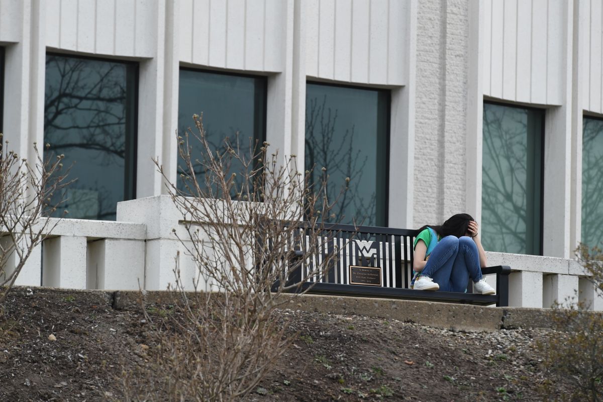 young person sits on bench outside large building, knees up on bench, face hidden behind them