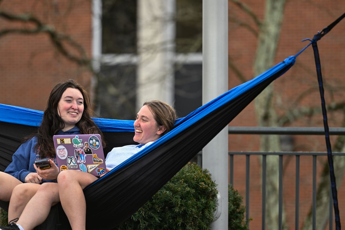 A blue and black hammock stretched out in front of a brick building holds two students. One is working on a laptop.
