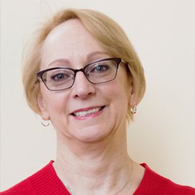 Headshot of Karen Kunz, WVU professor. Kunz is seated wearing an orange cardigan with a floral dress underneath. She has very short light-colored hair and wearing round glasses. 