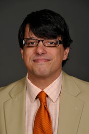 Headshot of man in tan suit, pink shirt and red tie with dark glasses on grey background