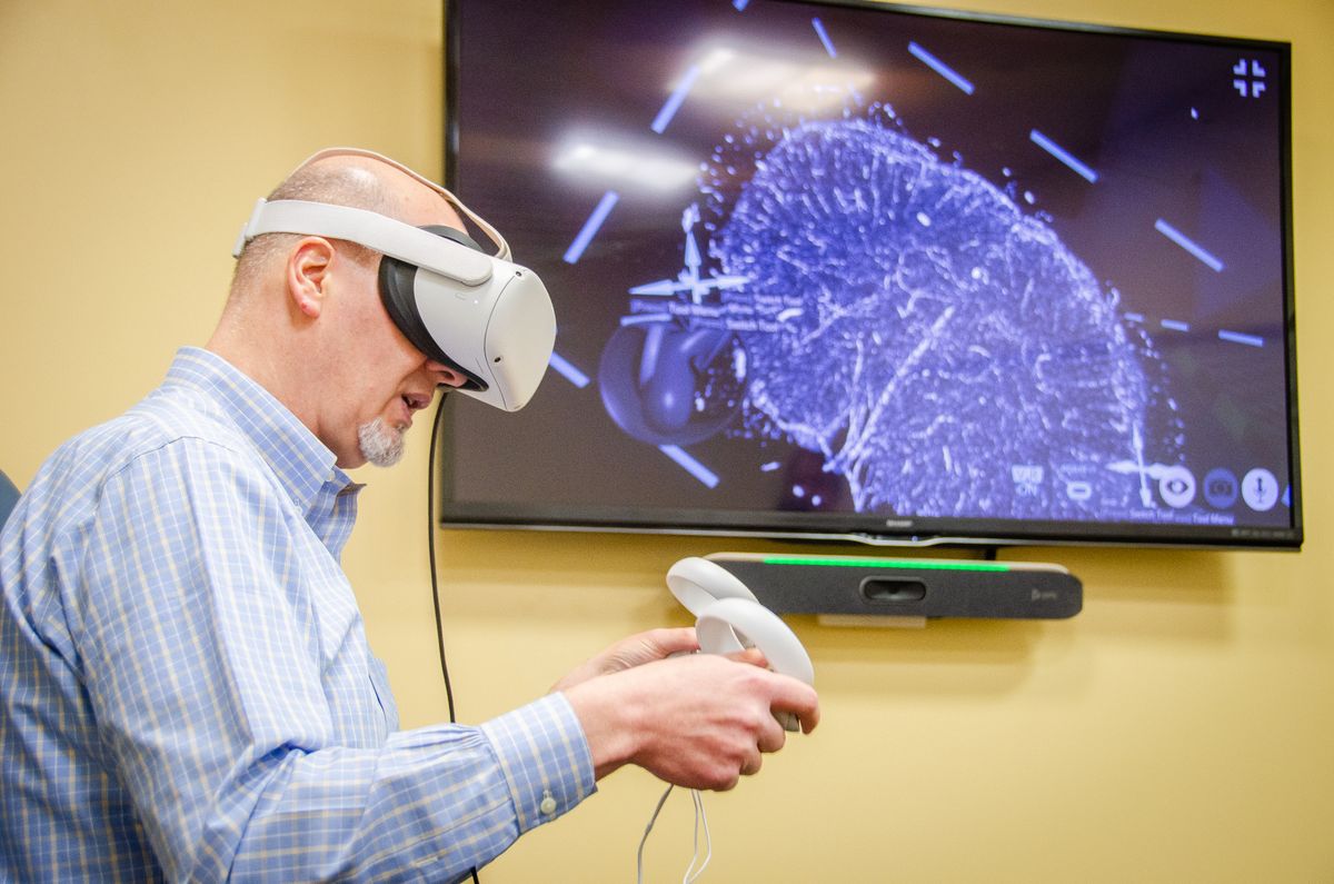 An image showing a WVU professor wearing a virtual reality headset and holding controllers while what he is seeing is displayed on a television screen on the wall to his left. 
