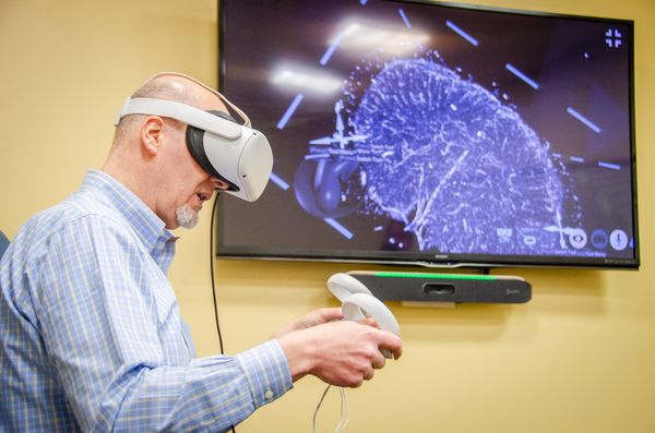 An image showing a WVU professor wearing a virtual reality headset and holding controllers while what he is seeing is displayed on a television screen on the wall to his left. 