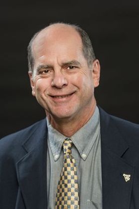 Headshot of WVU researcher Paul Ziemkiewicz. He is pictured in front of a dark background wearing a dark colored suit jacket over a gray shirt and a yellow and gray checked tie. He has a receding hairline and gray hair. 