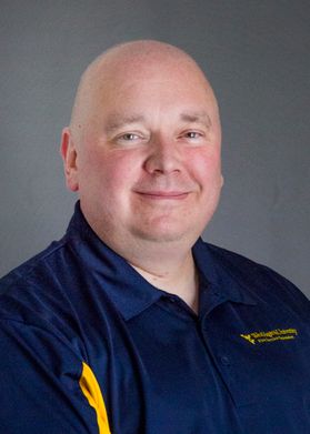 WVU Extension employee Mark Lambert poses for a headshot in front of a light gray wall. He is wearing a navy blue and gold WVU branded golf shirt. His head is shaved.