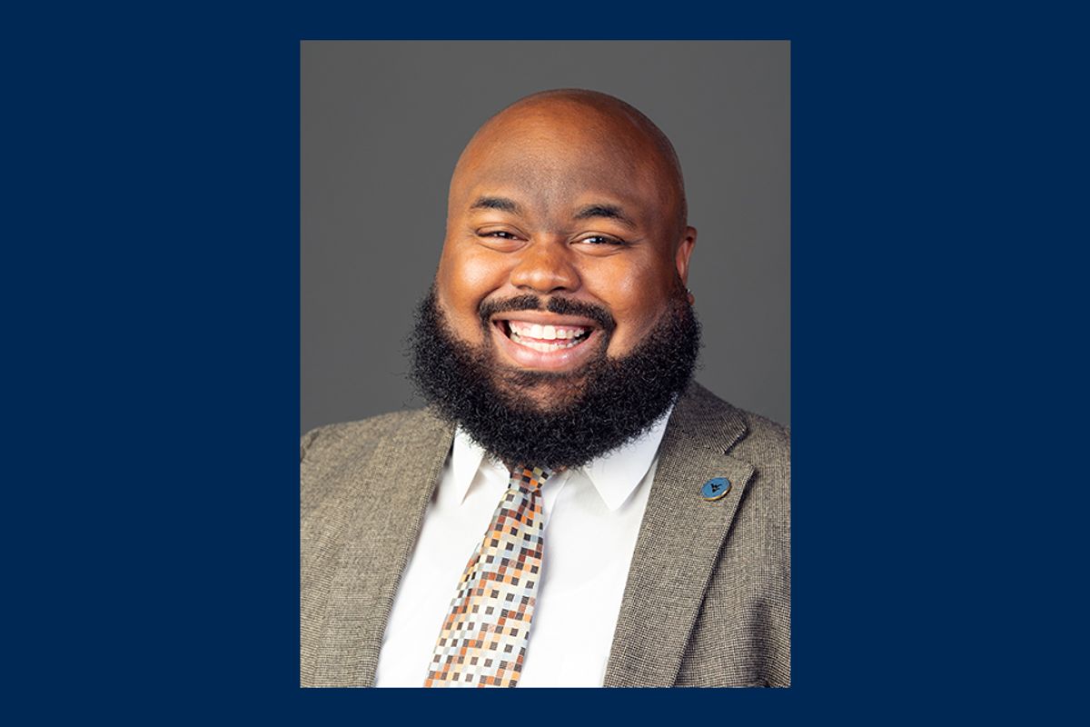A portait is in the middle of a blue background. Javier McCoy is smiling in front of a gray wall. Javier has a beard and is wearing a light gray suit jacket, gold tie with dots and white button-up shirt.
