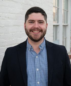 Headshot of WVU professor Chris Scroggins. he is pictured standing against a white brick building. He is wearing a dark colored jacket over a light blue shirt. He has short dark hair and a dark beard. 