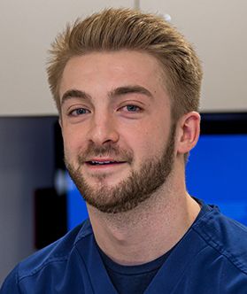 A young man with a beard smiles while wearing blue scrubs