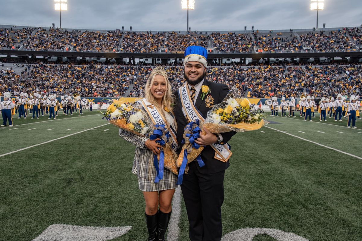 Two WVU students selected as Homecoming royalty pose together on the field wearing their crowns, sashes and holding bouquets of flowers. 