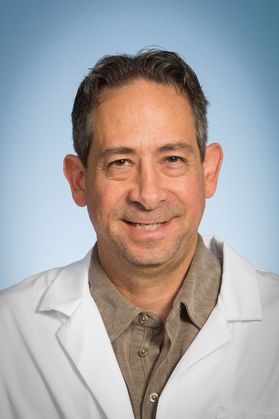 Headshot of WVU researcher John Hollander. He is pictured against a light blue background wearing a brown collared shirt under his white lab coat. He has short, dark hair. 