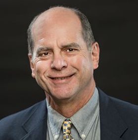 This is a portrait of Paul Ziemkiewicz who is wearing a dark blue jacket, gold and blue tie and blue button-up shirt while sitting in front of a black background.
