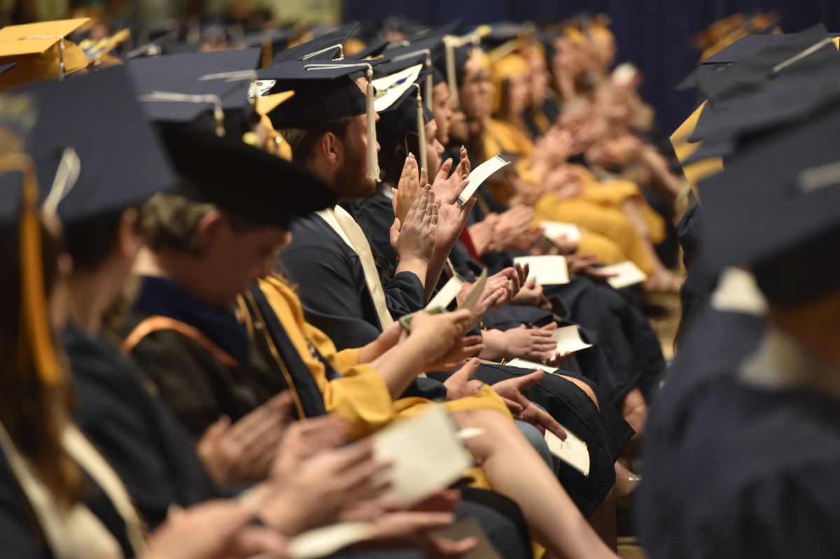 Generic photo of graduates seated in rows during a ceremony. The graduates are wearing black gowns with gold sashes. 