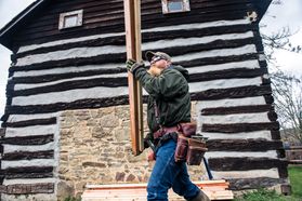 A man carries a beam in front of a log structure