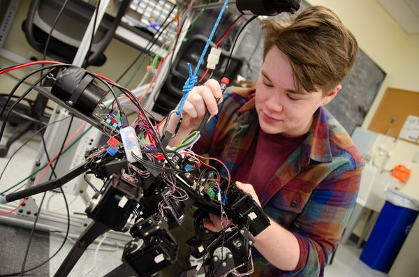 A student looks down while working on a black robot surrounded by wires.
