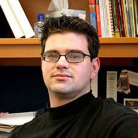 Headshot of WVU professor Brent McCusker. He is pictured in an office setting with books on shelves behind him. He has short black hair and black rimmed glasses. He is wearing a black shirt. 