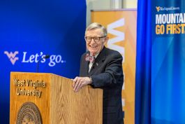 President Gordon Gee stands behind a wooden platform. He is wearing a dark suit and bow tie along with glasses. Behind him a blue banner includes white words that read, 'Let's go.'