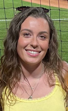Photo of WVU student Taylor Tetreault. She is standing in front of a baseball field and wearing a yellow tank top. She has long wavy brown hair and sunglasses sitting on top of her head. 