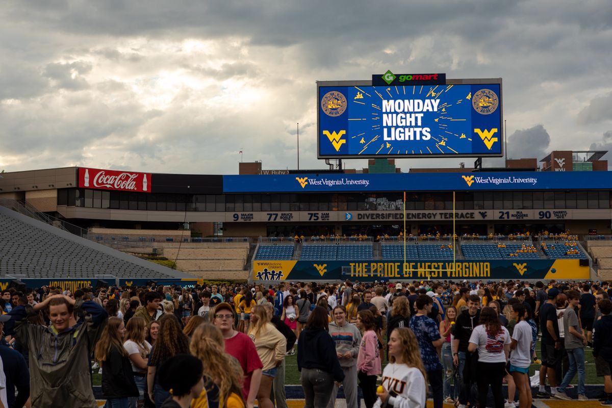 Monday Night Lights is shown in a graphic on the big screen at Milan Puskar Stadium against a cloudy sky while students congregate on the field.