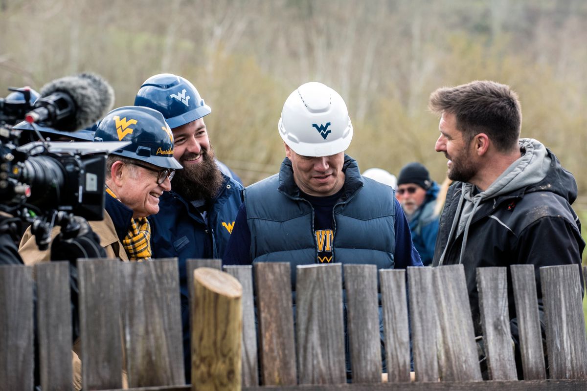Men in WVU hardhats stand behind a wooden fence. A video camera is to their right.