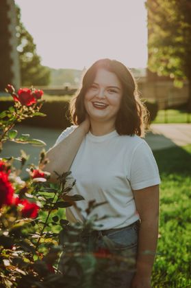 Photograph of scholarship recipient Adrianne Shimer. She is pictured outside with a flowering bush in the foreground. She is wearing a white T-shirt and has shoulder length brown hair. 