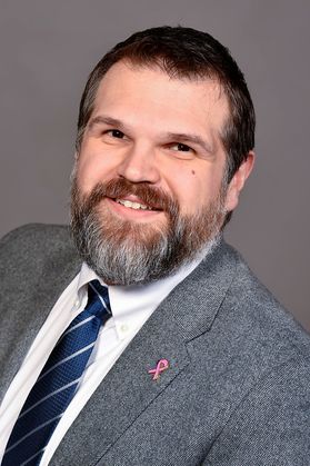 Headshot of George Zimmerman. He is smiling with brown hair and a full beard. He is wearing a gray jacket with a white dress shirt and blue striped tie. He is also wearing a pink, breast cancer lapel pin. 