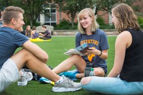 Three students sit on grass wearing grey WVU shirts looking at one another