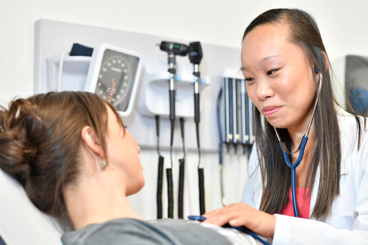 A female nurse practitioner of Asian descent with long dark hair exams a female patient in an exam room. There are medical tools hanging on the wall behind her and she is using a stethoscope during the exam. 