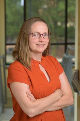 Headshot of WVU researcher Sarah Burke-Spolar. She is standing in front of a window with her arms crossed. She is wearing an orange, short sleeve blouse. She has shoulder length dark blonde hair. She is wearing glasses. 