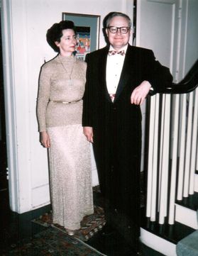 woman in beige sweater dress with gold belt and man in black suit with white button-up shirt and bow tie