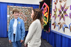 two women stand near hanging quilts