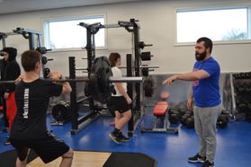A man in a blue t-shirt helps students with weight training