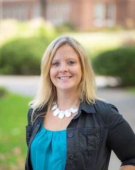 Headshot of WVU Professor Geah Pressgrove. She is standing outside with grass and bushes behind her. She has shoulder length blonde hair and is wearing a black jacket with a teal blue top. She is wearing a large necklace and is smiling in the photo. 