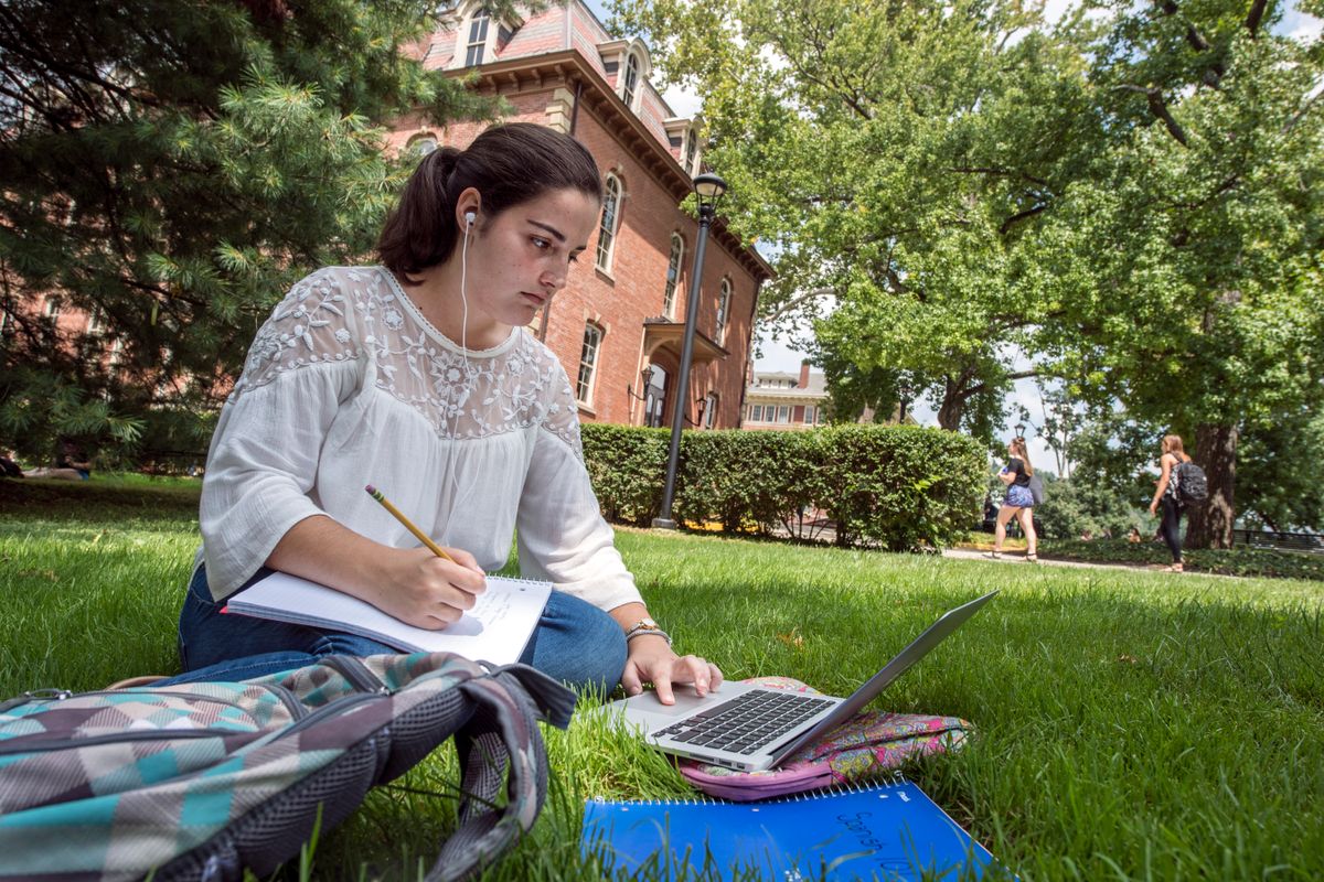 a young woman sits in the shade on the grass taking notes, looking at a laptop 