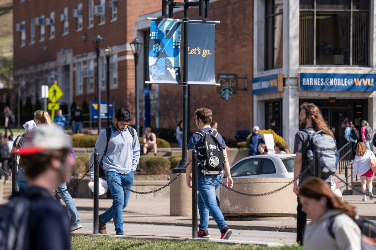 An image taken on the WVU campus. There are student walking throughout the photo. There is a car driving along the main road through campus in the frame. You can also see the WVU Bookstore in the distance. 