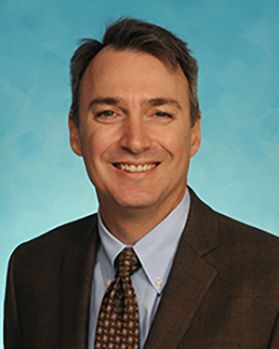 Headshot of WVU Medicine administrator Charles Mullett. He is pictured against a bright blue background and is wearing a brown suit over a light blue dress shirt and brown patterned tie. He has short, brown hair. 