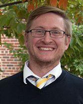 This is a headshot of Same Taylor. He is standing outside in front of a tree and is wearing black rimmed glasses, a block sweater, white button up shirt and gold and blue tie.