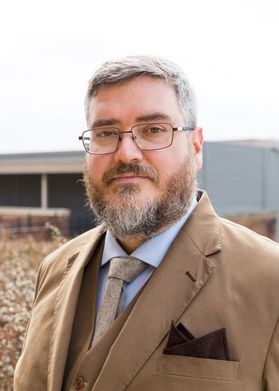 Headshot of WVU professor Sam Workman. He is pictured standing outside wearing a tan, three-piece suit with a light blue dress shirt and light colored tie. He has salt and pepper hair and a full beard. He also wears square framed glasses. 