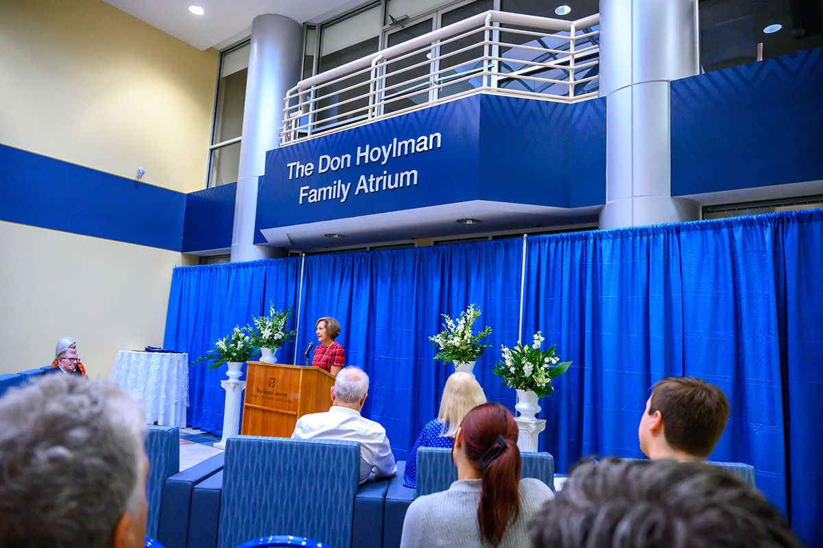 Donna Hoylman Peduto speaks at a podium in the atrium of the WVU School of Medicine to a crowd of people gathered for an event. 