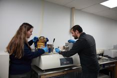 Two researchers work with lab equipment to study the nanostructure of a new oxide ceramic material. They are shown with their backs to the camera wearing blue surgical gloves and manipulating equipment. 