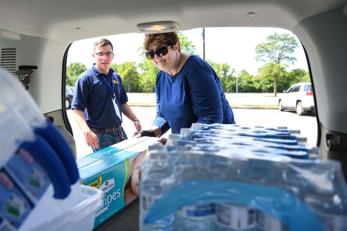 This the photo of the back of a vehicle. Bottled water is stacked in the forefront of the picture. Two people stand at the open back door. One is wearing a blue shirt with long sleeves and glasses. One is wearing a dark blue polo shirt with short sleeves.