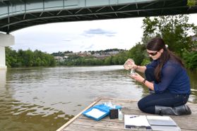 A WVU researcher tests water quality in the Monongalia River. She is kneeling on a dock near the water and pouring samples into test tubes. There is paperwork scattered around her. You can see the river and the very bottom of a bridge in the background. 