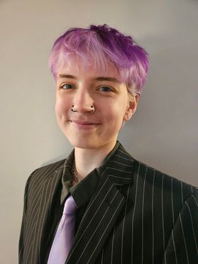 A portrait of Max Williams who is wearing a black striped suit with a purple tie and has short purple hair.