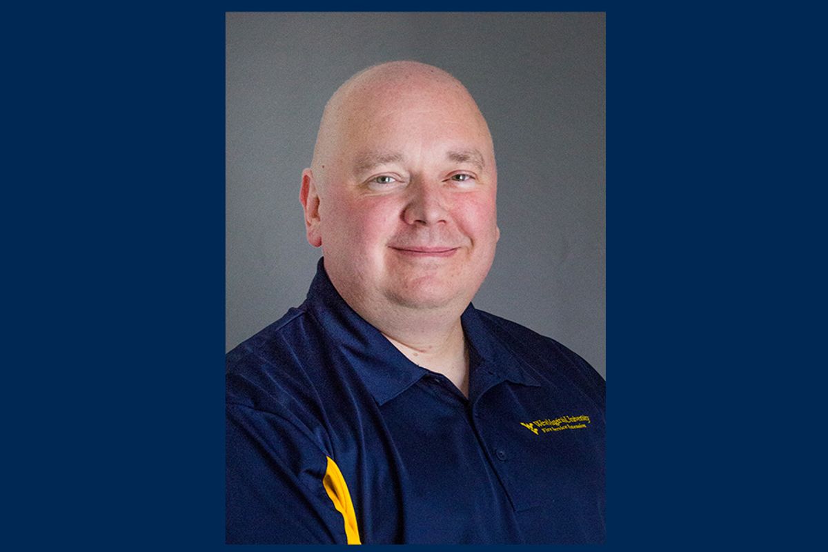This is a portrait of Mark Lambert on a blue background. Lambert is wearing a gold and navy blue polo shirt and is sitting in front of a gray backdrop.