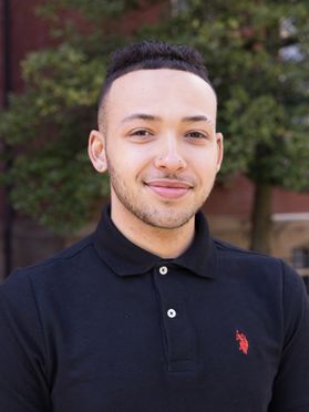 Headshot of WVU student Bryan Hill. He is pictured outside with a brick building and trees behind him. He is wearing a navy blue polo shirt buttoned to the top. He has short brown hair and a 5'clock shadow. 