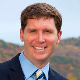 Headshot of WVU employee Greg Corio. He is pictured with colorful mountains behind him. He is wearing a navy blue sport coat with a light blue dress shirt and a gold and blue striped tie. He has light brown hair cut short. 