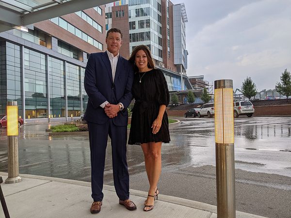 Smiling man in suit stands next to smiling woman in dress in front of WVU Hospital